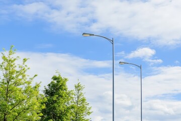 Two modern street lamps stand majestically against a bright blue sky, adorned with fluffy clouds, representing urban infrastructure and the essence of outdoor spaces and light.