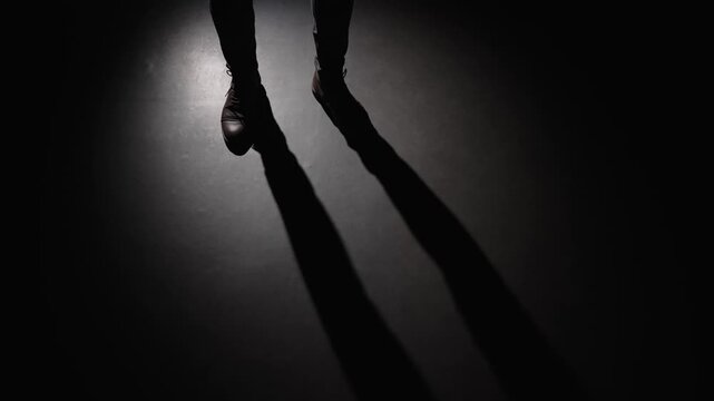 The silhouette of a man's legs entering a dark room. A mysterious  shadow stranger appears under the light of a lamp or lantern in the night, waiting for something.