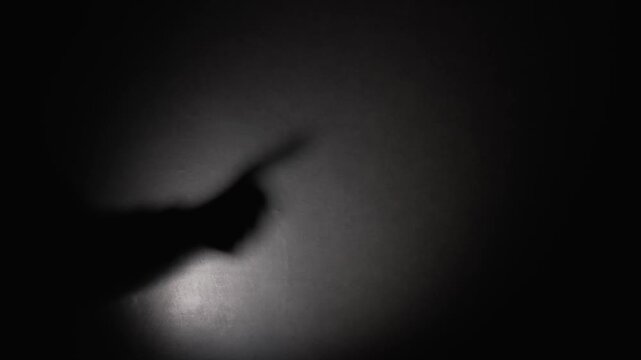 The shadow of a man's hand on a dark wall, giving a thumbs-up gesture against a black background. A mysterious stranger makes an approving gesture with his hand.