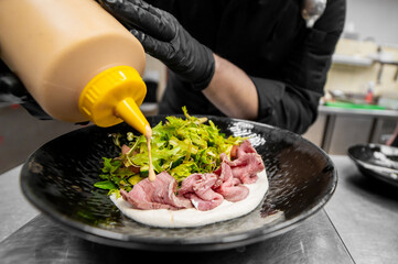 Chef in black gloves plates roast beef slices on a bao bun or salad base, drizzling a creamy sauce in a professional kitchen setting.