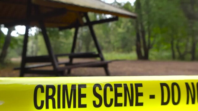 Close up view of a yellow colored crime scene forensic investigation police line ribbon in front of a camping area in a cloudy summer time forest with green trees and shallow depth of field, bokeh.