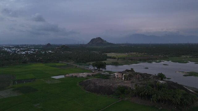 This atmospheric aerial footage captures a large wind farm set in a dynamic landscape. In the foreground, a rocky outcrop overlooks a large lake or reservoir with patches of vegetation.