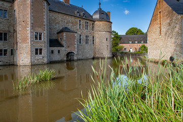 A scenic view of Château de Lavaux-Sainte-Anne in Belgium with its medieval walls rising above a peaceful moat, surrounded by greenery and bathed in warm sunlight under a clear blue sky.