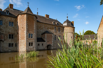 The tranquil moat of Château de Lavaux-Sainte-Anne reflects the historic stone walls and towers of this Belgian castle, surrounded by lush reeds and calm waters on a bright sunny day in the countrysid