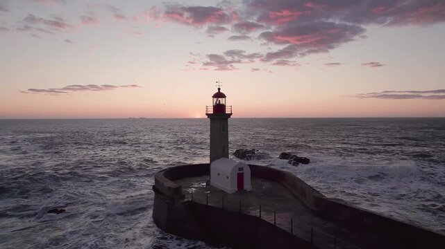 Drone orbits Farol da Barra lighthouse standing on rocky breakwater at Rio Douro estuary mouth during sunset, Atlantic waves surrounding fortified structure with horizon clouds glowing amber.