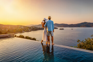 A romantic couple on summer vacation standing by the swimming pool enjoys a beautiful sunset behind the mediterranean sea