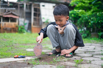 A young boy sits in the yard while digging soil using a small gardening shovel, playing with toy cars on the ground. The scene captures a candid outdoor moment of childhood play and creativity.