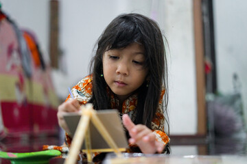 A young girl focuses while painting on a small canvas using a brush. She wears a colorful batik outfit as she works creatively at home, showcasing a genuine moment of childhood art and imagination.