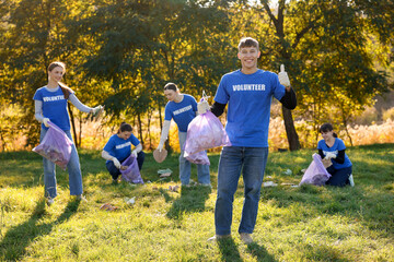 Young man showing thumbs up and group of happy volunteers with bags collecting trash in park