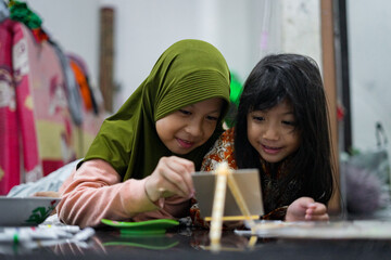 Two young girls painting together on a small easel while lying on the floor at home. They look focused and happy as they share a creative activity using brushes and bright colors.
