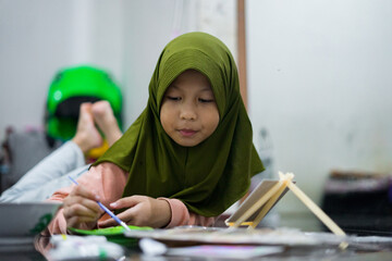 A young girl wearing a green hijab focused on painting indoors. She uses a brush and palette while sitting comfortably on the floor, expressing creativity through colorful artwork.