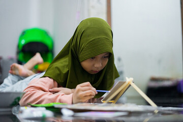 A young girl wearing a green hijab focused on painting indoors. She uses a brush and palette while sitting comfortably on the floor, expressing creativity through colorful artwork.