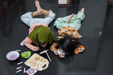 Two young girls are lying on the floor while painting and coloring. The girl in the green hijab focuses on her canvas, while the other girl smiles and watches closely. 