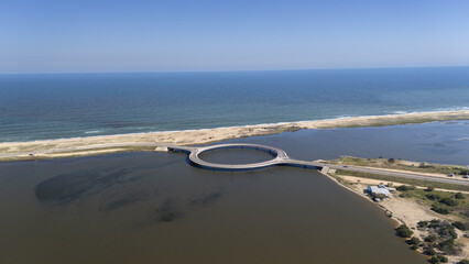 Aerial view of a circular bridge spanning tranquil waters where the lagoon meets the ocean's edge, a serene blend of blue and earthy tones, Faro de José Ignacio, Departamento de Maldonado, Uruguay.
