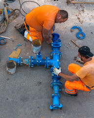 Two construction workers in orange workwear are assembling a large blue water pipe system on...
