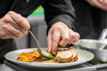 A close-up shot of a chef's hands using a spoon to add sauce to a plated gourmet dish of sliced pork tenderloin, roasted potatoes, and peas in a professional kitchen setting.