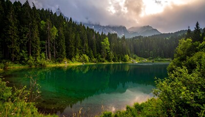 Still teal lake surrounded by forest, with distant mountains under a cloudy sky