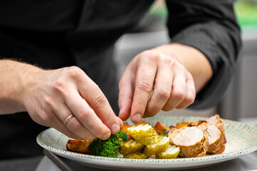 A chef adds a final garnish of parsley to a plate of sliced pork tenderloin with pickles and roasted vegetables in a professional kitchen.