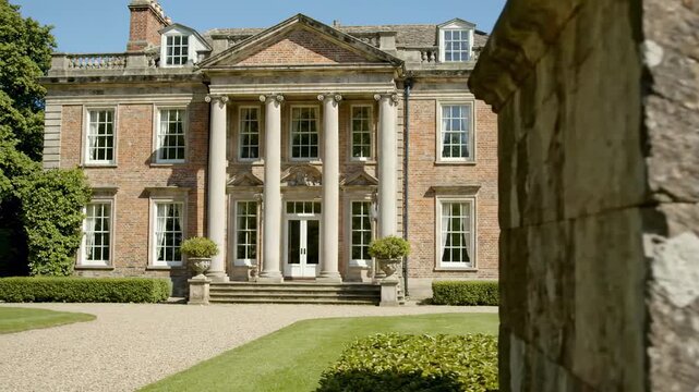 A brick manor with a classical portico of white columns and a pediment, flanked by manicured lawns and a gravel path; a weathered wooden foreground frames the polished fa&ccedil;ade - georgian home