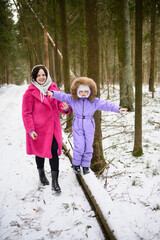 Mother supports child as she walks along snowy path on fallen tree, protecting, holding, and encouraging her. Mother helps girl balance on mossy log on snowy path. Winter walks in snowy forest.