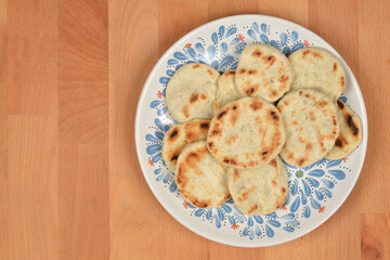 A collection of small, round, toasted flatbreads with golden-brown grill marks is displayed on a white and blue decorative plate resting on a wooden surface