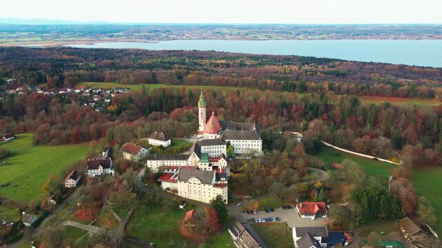 Aerial view of Andechs Abbey, a monastery atop a hill surrounded by autumn foliage, contrasting with the distant lake, Andechs, Bayern, Germany.
