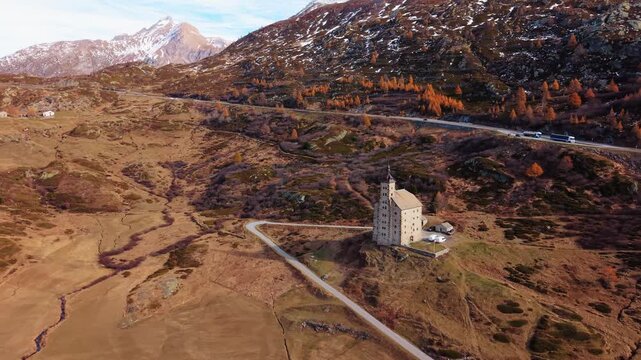 Aerial view of Simplon Hospice standing tall on a hill, with road traffic nearby, amidst autumnal colors and snow-capped mountains, Simplon Hospice, Wallis, Switzerland.