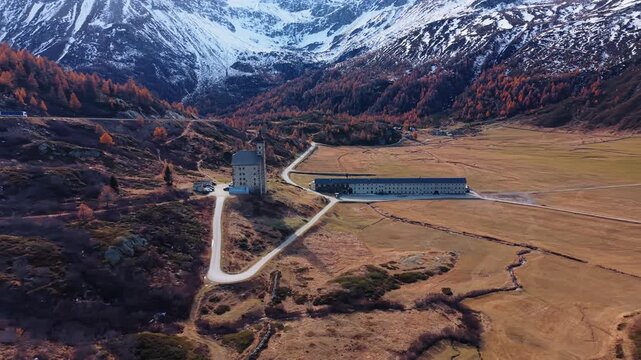 Aerial view of Simplon Hospice's tower and long building, contrasting with the golden fields and snow-capped mountains, Simplon Hospice, Wallis, Switzerland.