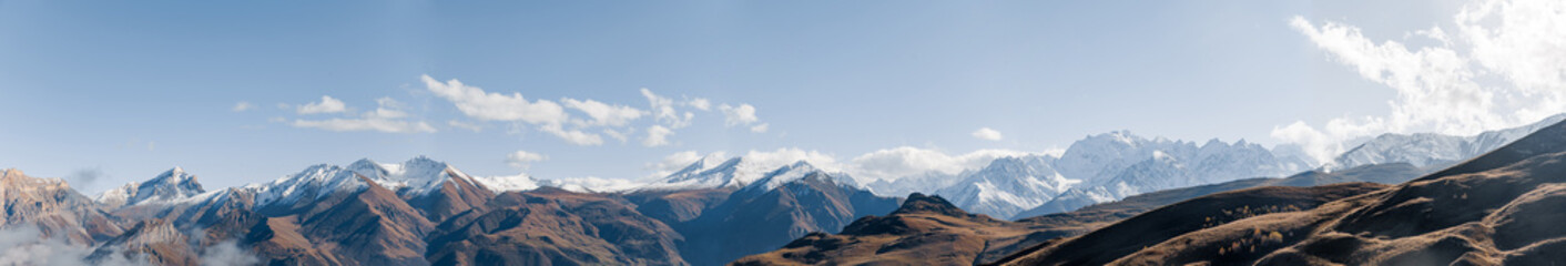 Panoramic view of the majestic Caucasus mountains in Kabardino-Balkaria. Snow-capped peaks under a clear blue sky. Rugged terrain and rocky slopes visible.