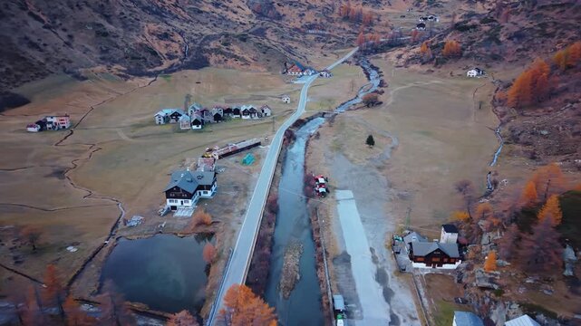 Aerial view of houses with dark roofs, a pond, road, and river creating a serene landscape with mountains in the background, Formazza, Piedmont, Italy.