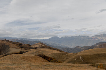 Panoramic view of the mountainous landscape in Kabardino-Balkaria. Rolling hills with dry grass and distant snow-capped peaks under a cloudy sky.