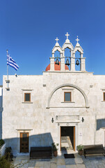 The Panagia Tourliani Greek Orthodox monastery in Mykonos with whitewashed walls, a red dome, an ornate bell tower, and a prominently displayed Greek flag