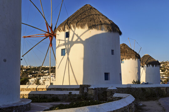 
Group of traditional windmills in Mykonos, Greece, with white cylindrical structures and conical thatched roofs, set on a stone platform under sunrise sky - Powered by Adobe