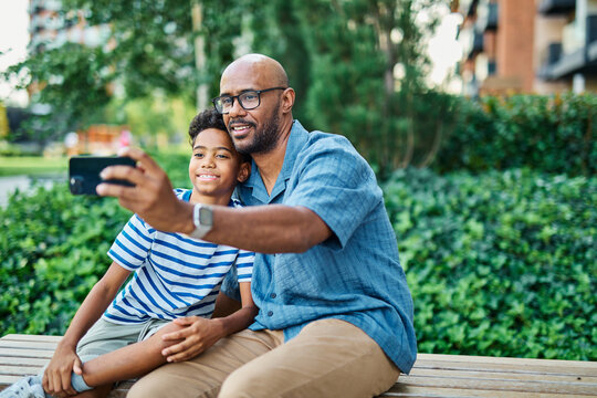 Portrait of father and son having fun using a mobile phone and taking a selfie photo with camera outdoors in park or nature,, family life, parenting, love and bonding concepts - Powered by Adobe