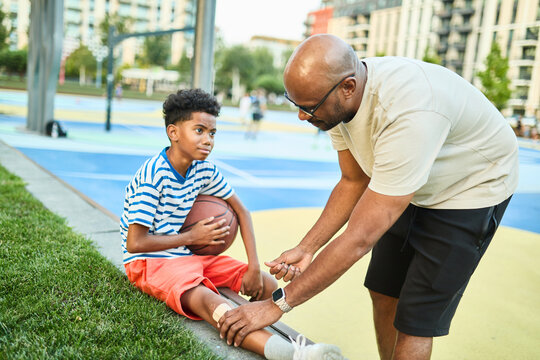 African american father and boy smiling, standing together playing basketball and sitting injured father helping and supporting son or coach on a colorful basketball court