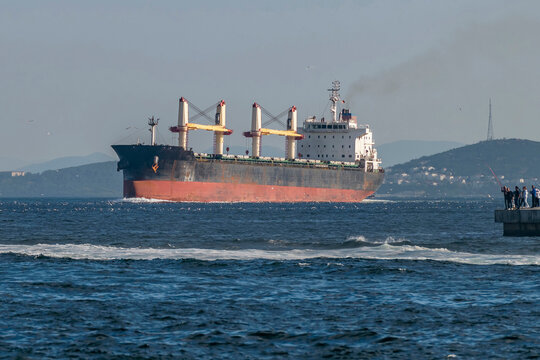 A large bulk carrier vessel sails on the Bosphorus, Istanbul, T&uuml;rkiye, passing near a pier where some fishermen can be seen