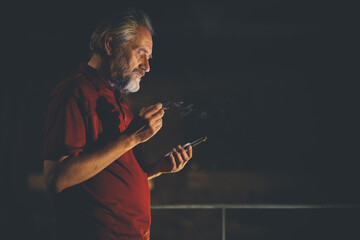 Older man in red shirt waiting for something and using mobile phone on night city location