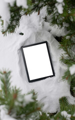 Vertical mockup photo of a digital tablet with blank white screen lying in fresh snow among evergreen branches, winter flat lay background for holiday sales, apps, ebooks or social media design.