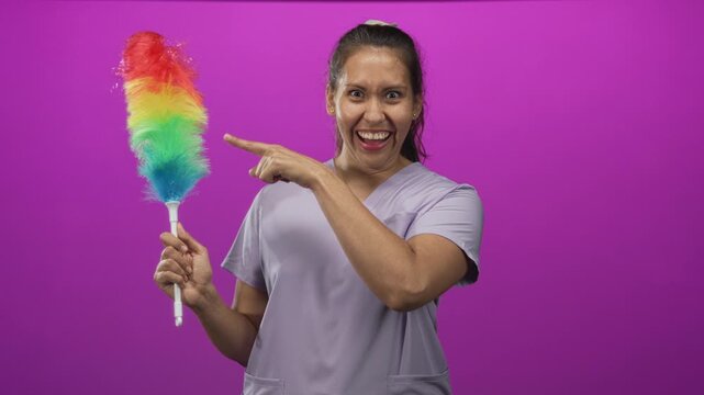 Young hispanic woman cleaner holding rainbow feather duster and pointing finger in purple studio, wearing lavender scrub top and smiling broadly; playful.