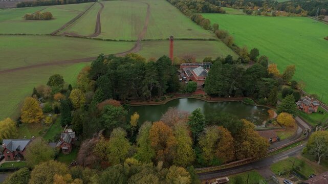 Aerial view of a water tower and trees with autumnal hues surrounding a pond reflecting the overcast sky, contrasting with green fields, Nottingham, England, United Kingdom.