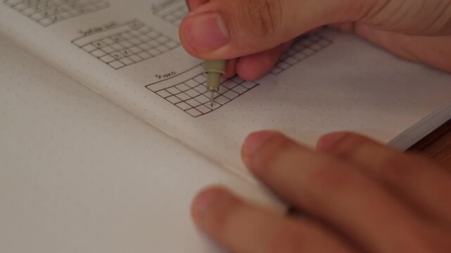 Close-up static shot of a hand and pen filling out habit tracker grid inside journal spreadsheet.