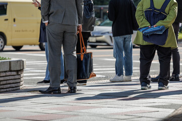 Businessman holding casual long handled tote bag stands with diverse of casually dressed pedestrians on a chilly morning, waiting for city crosswalk traffic light to change.