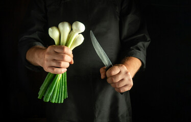 A skilled chef holds a bunch of green onions in one hand and a sharp knife in the other, ready to chop for a flavorful dish in a dark kitchen