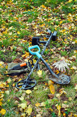 Various metal detecting tools are set on the ground filled with colorful fallen leaves in a park. The scene captures the essence of autumn and outdoor exploration