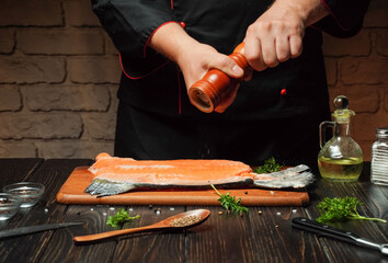 A chef in a black jacket is sprinkling pepper over a fresh salmon fillet resting on a wooden cutting board in a cozy kitchen setup with herbs nearby
