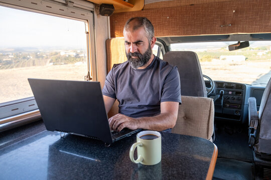 A middle-aged bearded man types on his laptop inside a camper van, with natural light entering through the window and a mug of coffee on the table in front of him.