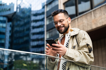 Young professional in a modern business district checks his phone—smiling at good news and managing messages, payments, maps, and schedules on the go while commuting between meetings across the city