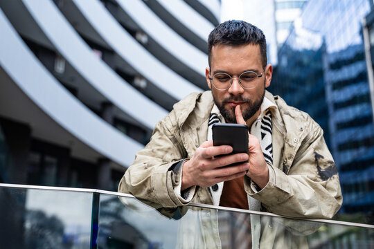 Young professional in a modern business district checks his phone—smiling at good news and managing messages, payments, maps, and schedules on the go while commuting between meetings across the city