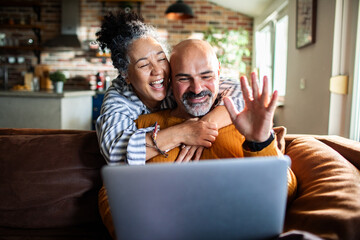 Happy senior couple hugging and waving on video call at home