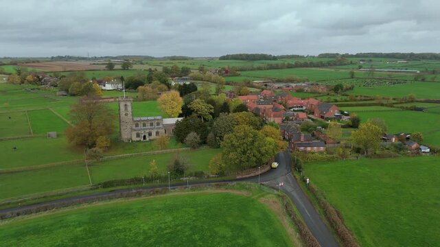 Aerial view of a church and village surrounded by green fields, a road cutting through the landscape under a cloudy sky near Loughborough, England, United Kingdom.